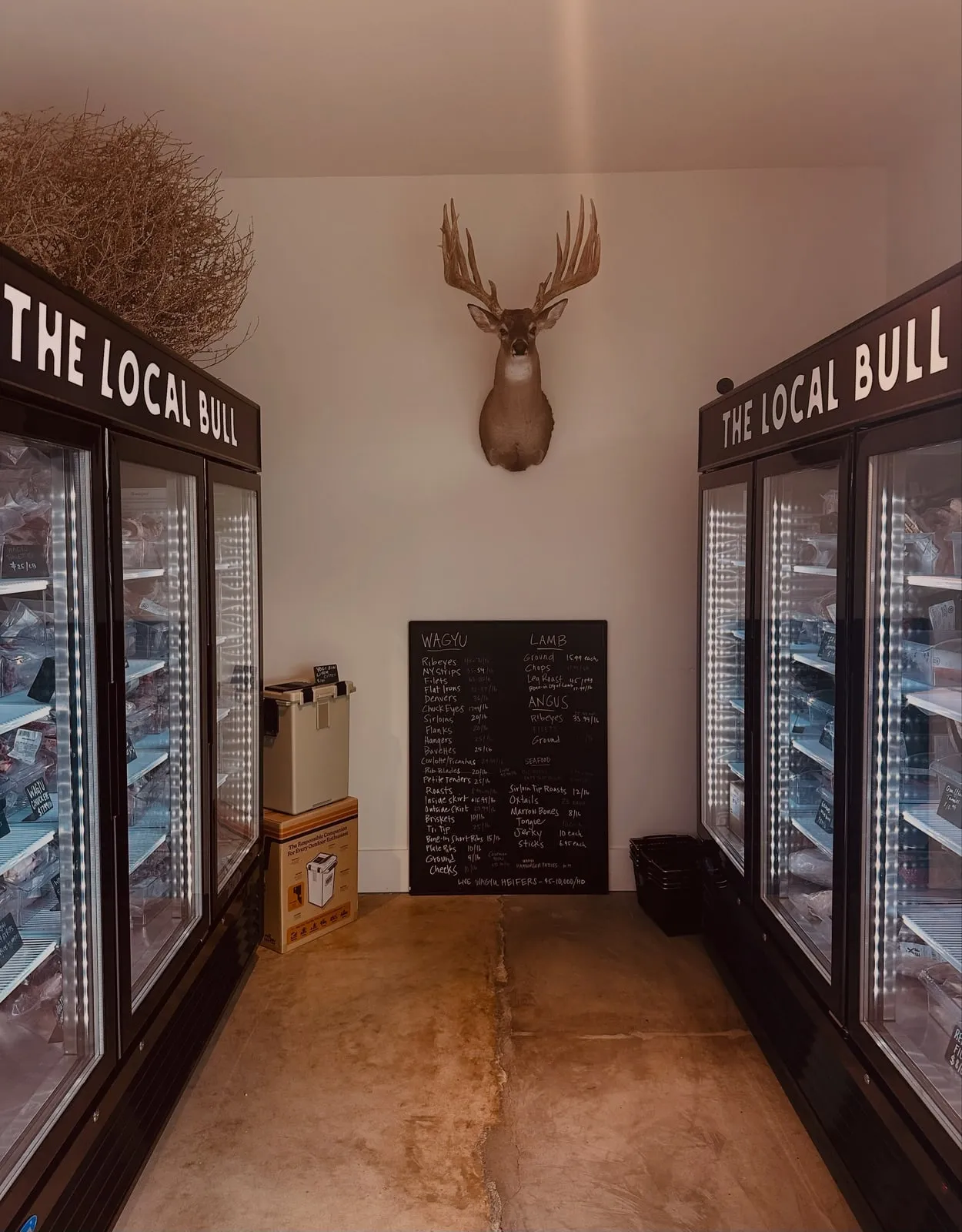 Inside The Local Bull: two freezer cases of Wagyu under a mounted whitetail, chalkboard menu on the back wall